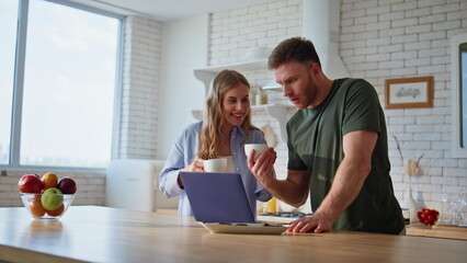 Wife bringing tea husband looking laptop at kitchen. Couple enjoying beverage