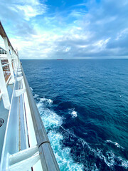 View of the side of a cruise ship in the open sea with waves crashing against its side in a deep blue tone with a cloudy sky on a summer day. Mention of family vacations and relaxation.