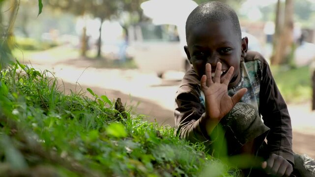Portrait of innocent kid waving at the camera in an African village. Small huts among trees, people on the streets, green forests, and everyday life capture the simplicity of African village living.