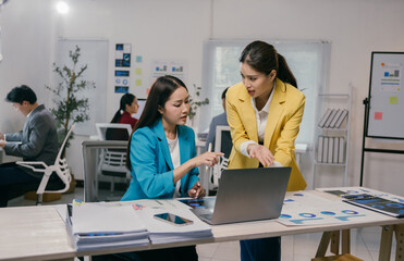 Two asian businesswomen are discussing a project using a laptop in a busy office, analyzing charts and collaborating on a task