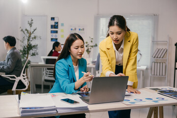 Two asian businesswomen are discussing over a laptop analyzing charts and reports pointing at the screen collaborating on a project in a busy office