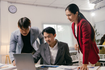 Three asian businesspeople are collaborating on a project, gathered around a laptop in a modern office meeting room, analyzing data and discussing strategies