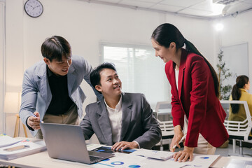 Three asian businesspeople are discussing work, using a laptop and documents, in a modern office environment, collaborating on a project and analyzing financial data