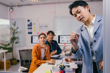 Asian businessman is drawing a business plan on a transparent whiteboard during a corporate meeting, while his colleagues are sitting at the table and looking at him