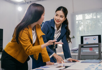 Two asian businesswomen are discussing and brainstorming about new business project, pointing at charts and graphs placed on table in meeting room