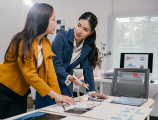 Two businesswomen leaning over a table covered with financial charts and graphs, actively discussing data and collaborating on a project in a modern office setting