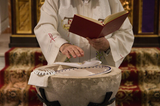 The priest blesses with his hands the water of the baptismal font to proceed to the sacrament of baptism in a catholic church. On the edge of the font the silver shell and the white handkerchief.