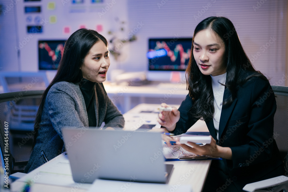 Poster two young businesswomen are working late in the office, collaborating on a project using a laptop an