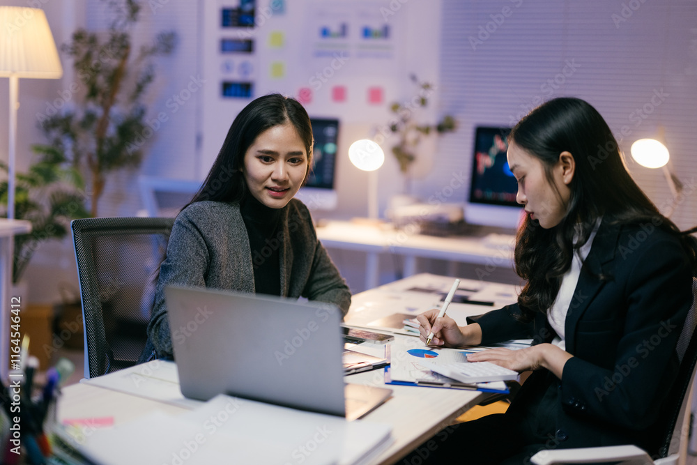 Poster two young asian businesswomen are working late in a modern office, collaborating on a project using 