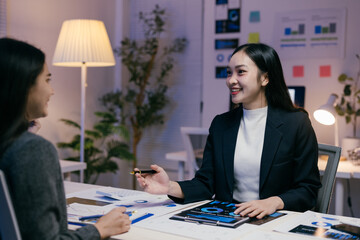 Two young businesswomen are sitting at a desk in a modern office, working late at night, discussing marketing strategy using charts and graphs, smiling and collaborating effectively