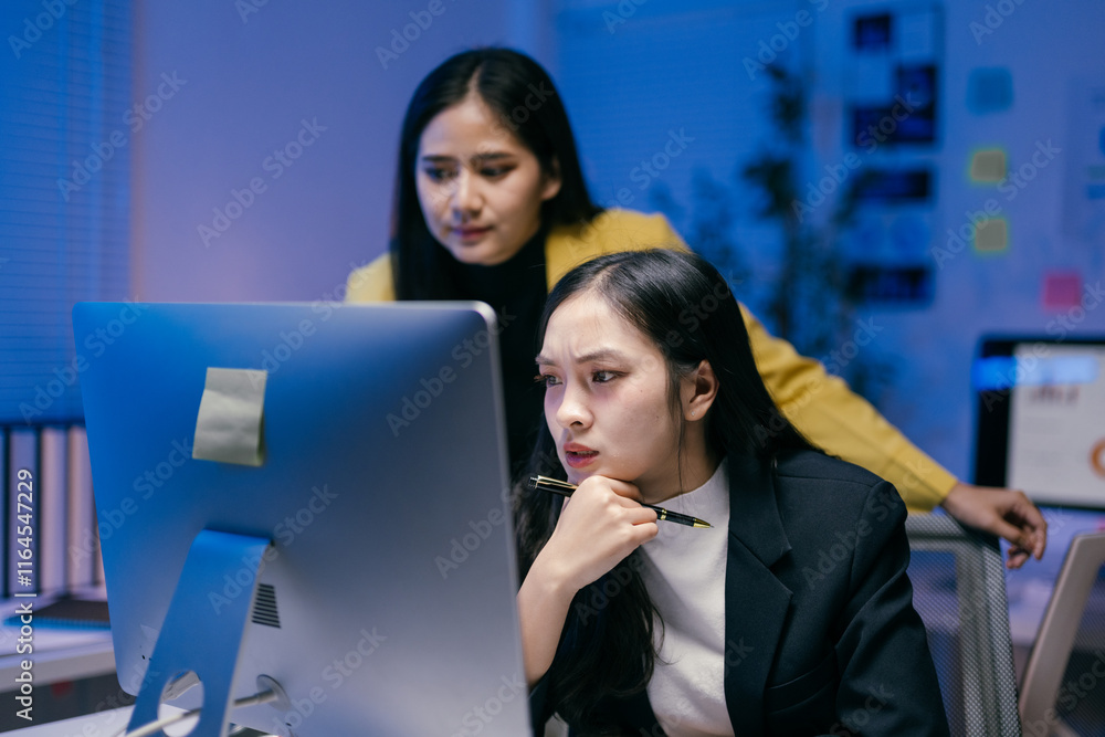 Aufkleber Two asian businesswomen working late in office using desktop computer and experiencing issues with software while one of them is holding a pen and thinking about solution