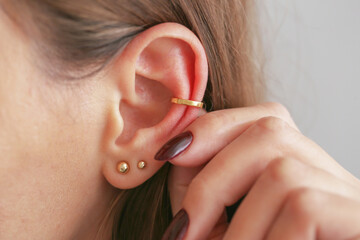 Golden ear cuff jewelry on irritated red ear. Highlight of putting on fashionable earring accessory. Elegant ear decoration closeup. Ear infection background.