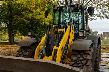 A large yellow and black loader is clearing fallen leaves in a park