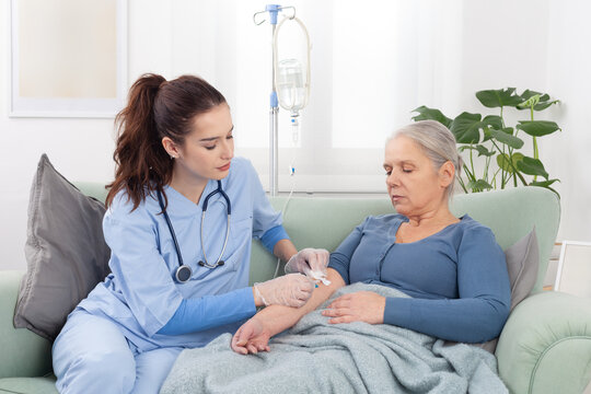 Young friendly nurse or doctor help sick senior elderly woman patient in receiving IV drip infusion and vitamin injection therapy sitting on sofa in medical clinic or at home. Health care concept