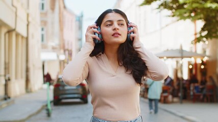 Happy overjoyed Indian young woman in wireless headphones dancing listening favorite energetic disco music in smartphone dancing outdoors. Hispanic girl walking on urban city street. Town lifestyles