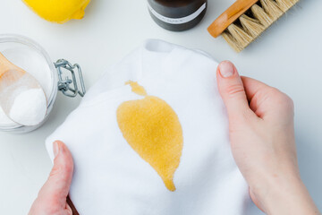 Close up a person holding white fabric with a stained yellow in the background of baking soda and a lemon, showcasing a DIY cleaning method using natural products to remove stains. top view