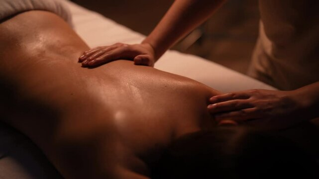 Masseuse gently applying massage oil on woman back lying on massage table, providing relaxing and rejuvenating spa treatment in beauty parlor with dark interior, close-up. Shooting in slow motion.