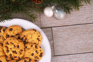 Top view of chocolate chip cookies on a plate and a Christmas tree branch displayed a white wooden plank in background. Copy space for your text. Christmas holiday concept.