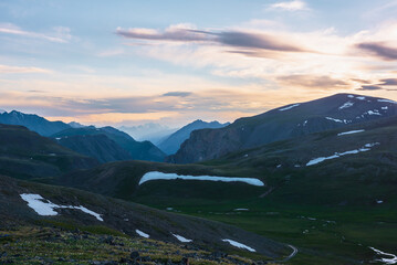 Scenic view from flowering grassy stony hillside to alpine valley, green pass, deep gorge and big sharp mountain peak silhouette against large snowy top far away under vivid sunset color cloudy sky.