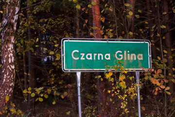 Tablica z nazwą miejscowości CZARNA GLINA w lesie jesienią po zmroku. Tablica na początku wsi  położonej wśród lasów, w anturażu  jesiennym o zmierzchu. 