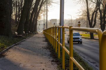 Żółte stalowe bariery wzdłuż chodnika położonego wyżej niż droga w mglisty jesienny dzień. Zdjęcie jesiennego dnia w mieście (Ostrowiec). Chodnik biegnący przez zbocze niewielkiego wzgórza.