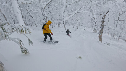 Couple of snowboarders enjoy riding through a snowy forest, surrounded by bare trees blanketed in...