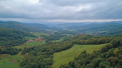 Obraz premium High angle view of a valley nestled between forested hills. Lush greenery, fields, and a village are visible. Cloudy sky.