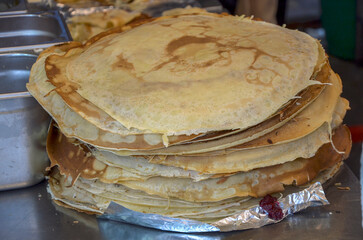 Large stack of thin, golden-brown crepes sits on a kitchen counter, ready to be served. The crepes...