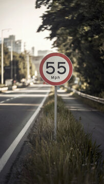 Scenic Roadside Speed Limit Sign, 55 MPH, with Urban Background, Asphalt Highway, Greenery, and a Serene Morning Light Effect