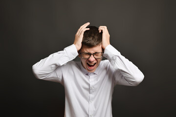 a young man with glasses, holding his head in his hands, with his eyes closed, screaming in pain and anger. Studio photo, on a gray background