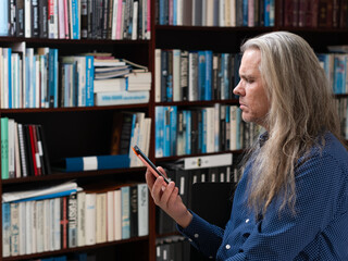 Side view of a man with long hair reading on his e-book in front of a bookshelf