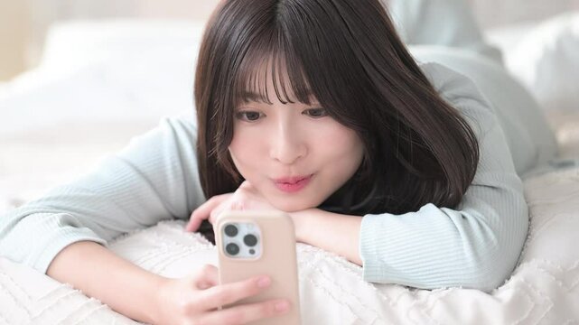 ベットで寝転がってスマホを見る笑顔の若いかわいい女性のクローズアップの動画