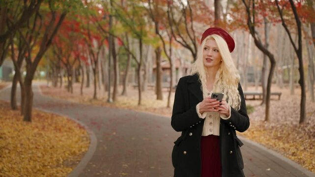 Blonde European woman using her phone texting as she walks on path surrounded by autumn leaves, slow motion, soft smile with wonder and admiration