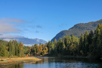 A scenic mountain valley with lush green forests, a pile of logs in the foreground, and a vibrant blue sky with dramatic clouds.