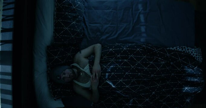 A man lies awake in bed, illuminated by faint moonlight streaming through window blinds. Wearing a beanie, he appears lost in thought reaching across to a space on the bed he wants filled by a person