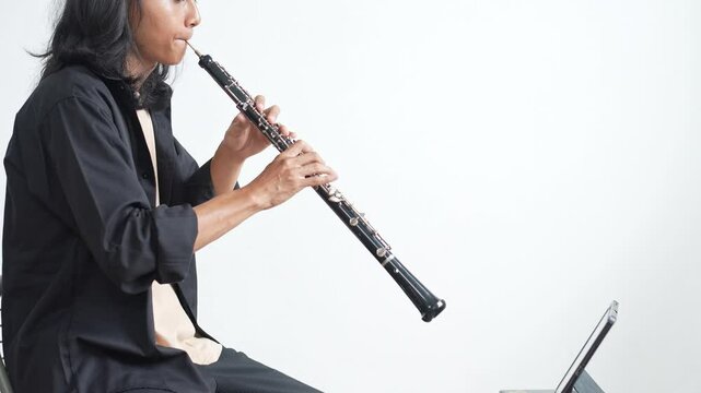 Asian Man Musician Sitting On A Chair Playing Oboe While Looking At Video Tutorial Online On Tablet