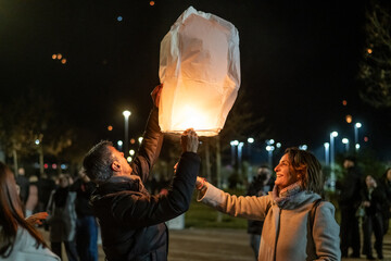 Couple releasing glowing sky lantern during nighttime festival, joining crowd releasing floating lights, symbolizing shared hope and collective celebration