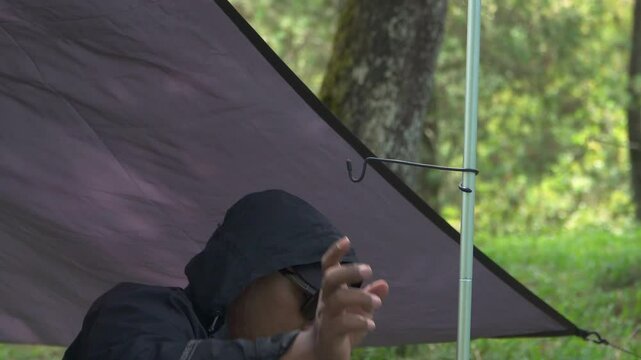 A solo traveler is preparing a flysheet tent in a green and lush pine forest area. Asian man adventuring alone in the jungle and setting up a trap tent from flysheet for bushcraft and camping