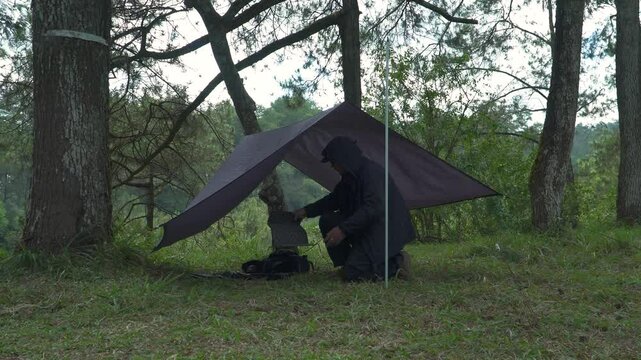 A solo traveler is preparing a flysheet tent in a green and lush pine forest area. Asian man adventuring alone in the jungle and setting up a trap tent from flysheet for bushcraft and camping