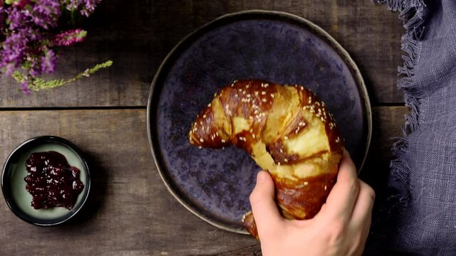 Overhead shot of a hand reaching for a golden pretzel croissant on a rustic blue plate. A bowl of berry jam and a textured wooden table with decorative purple flowers complete the cozy breakfast scene