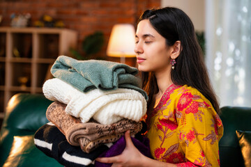 Indian woman in kurta folding woolen clothes, enjoying the fresh fragrance while relaxing on the sofa