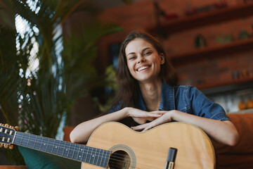 Woman sitting on couch with acoustic guitar in front of potted plant in cozy home setting