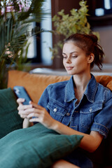 Obraz premium Woman relaxing on couch, wearing denim shirt and blue jeans, browsing on smartphone