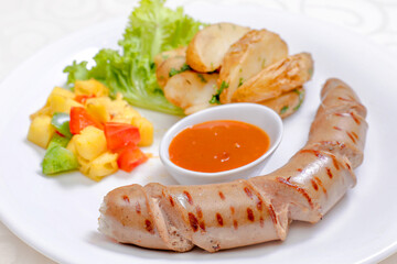 A grilled sausage served with potato wedges, a side salad, and a bowl of chili sauce in isolated white background
