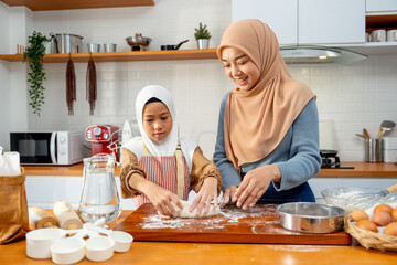Wide shot of young Asian muslim mother help and teach her daughter to threshing flour by hands on wooden tray for making the bakery in the kitchen of their house.