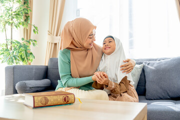 Asian muslim mother sit on sofa beside of her daughter and hug her child they also look face each other with smiling in concept of happiness muslim family at home.