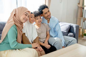 Fototapeta premium Asian muslim family compose of young mother, father, daughter and son hug and stay together on sofa in living room of their house and look forward and left direction of image with smiling.