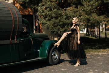 woman standing next to a green retro car. She is dressed in a long black dress with transparent...