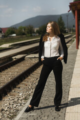 The woman stands in a relaxed position, with her hands in her trouser pockets, and looks to the side, eyes slightly squinted. Train tracks, greenery and mountains are visible in the background.