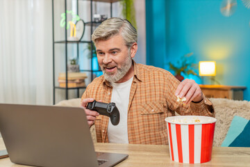 Caucasian mature man playing video games on laptop with gamepad joystick. Middle-aged guy sitting...